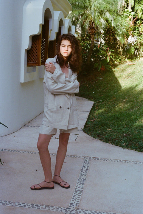 Brunette woman standing in the full Easy Going Linen Suit in Sand with black sandals. She has her arms wrapped around herself.