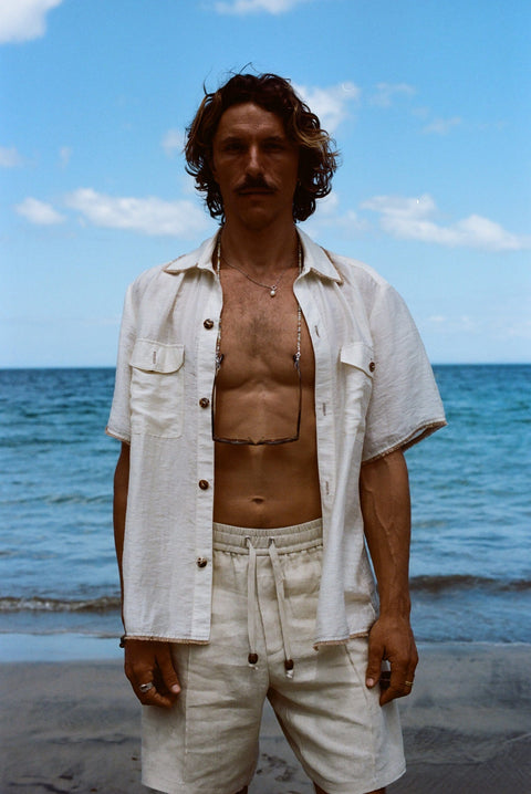 Brunette man standing on beach while wearing the sand linen Easy Going Shorts with our featherweight Resort Shirt fully unbuttoned.