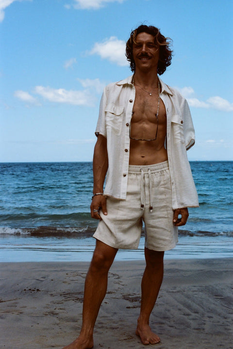 Brunette man standing on beach while wearing the sand linen Easy Going Shorts with our featherweight Resort Shirt fully unbuttoned.