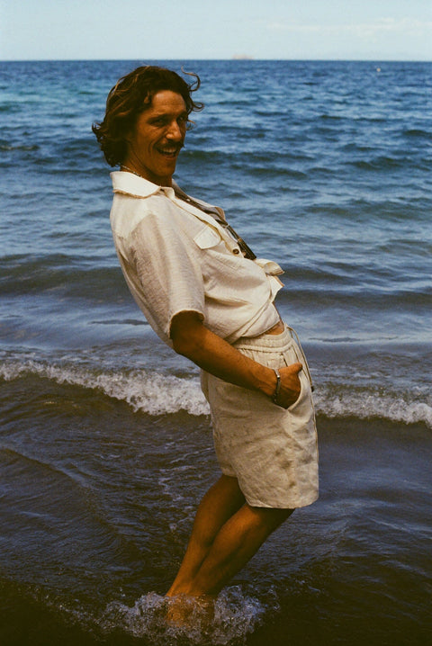 Brunette man standing in the ocean, wearing the sand linen Easy Going Shorts and our featherweight Resort Shirt and leaning backwards, with his hands in his pocket. 