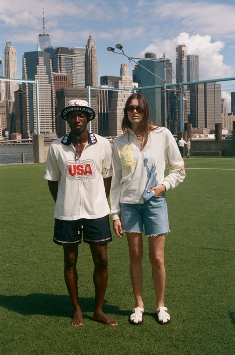 Two models wearing FIFA collection tops stand on a grassy field with the New York City skyline behind them.
