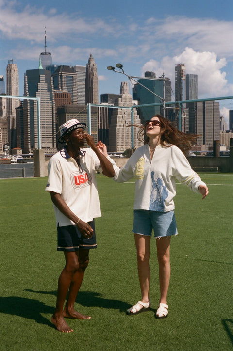 Two models wearing FIFA collection tops stand on a grassy field with the New York City skyline in the background. One wears a cream polo shirt with a red USA graphic, and the other wears a cream terry quarter-zip pullover with yellow FIFA embroidery and a blue Statue of Liberty graphic.