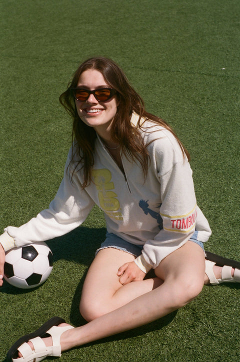 Model seated on a grassy field beside a soccer ball, wearing a cream terry quarter-zip pullover with FIFA and Statue of Liberty graphics.