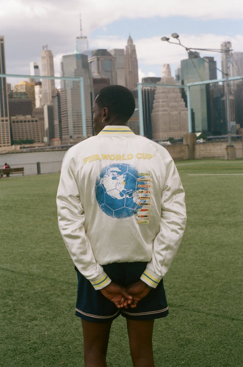 Person wearing a white jacket with a world cup design, standing on a grassy field with a city skyline in the background.