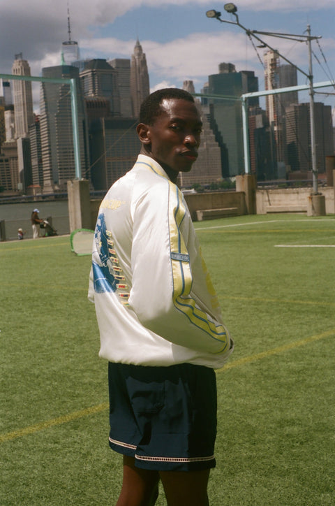 Person wearing a white jacket with a world cup design, standing on a grassy field with a city skyline in the background.
