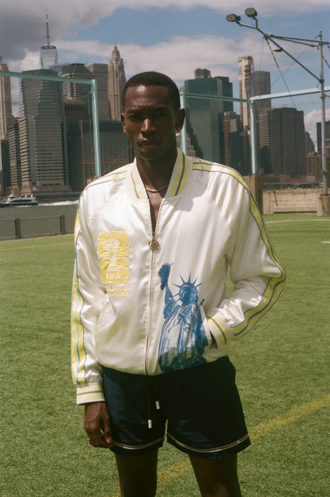 Person wearing a white jacket with a graphic statue of liberty on the front on a sports field with city skyline in the background