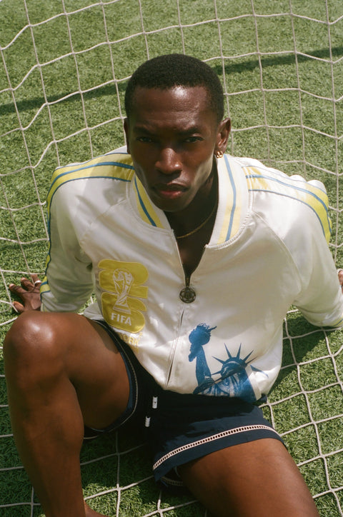 Man wearing FIFA Souvenir Jacket and swim trunks, sitting on a soccer net on a field. 