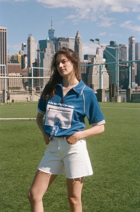 Model wearing a blue terry quarter-zip polo with FIFA World Cup embroidery and a mesh Statue of Liberty pocket on a soccer field.