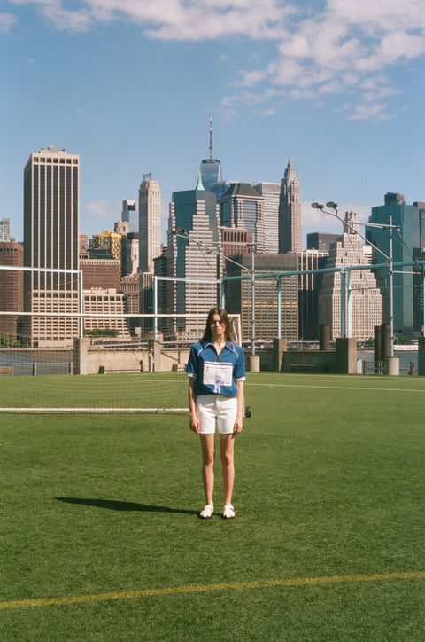Wide shot of a model wearing a blue terry quarter-zip polo with FIFA World Cup embroidery, standing on a soccer field with the New York City skyline behind them.