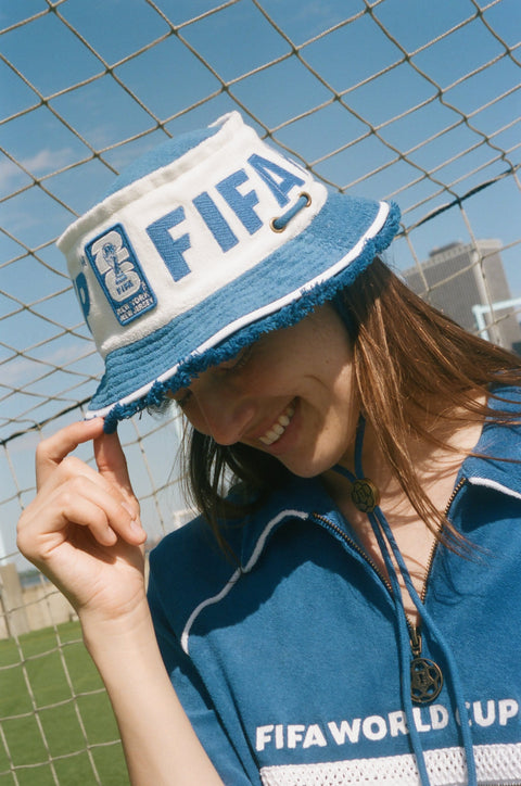 Close-up of a white-and-blue FIFA bucket hat worn with a blue terry quarter-zip polo featuring FIFA World Cup 26 embroidery.