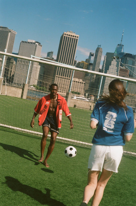 Two models playing soccer on a field, wearing FIFA collection tops in front of a goal net and the New York City skyline.