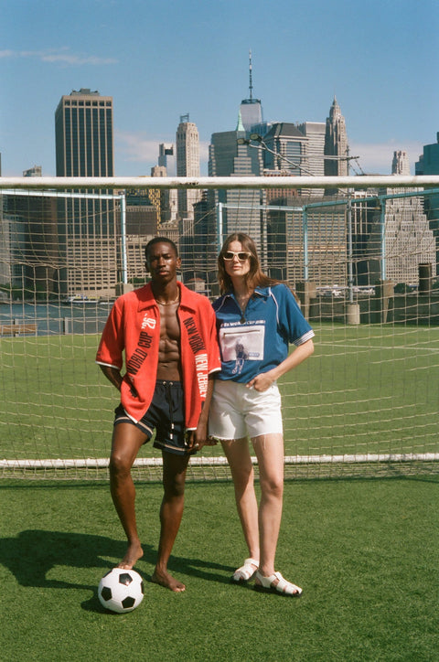 Two models wearing FIFA collection tops stand in front of a soccer goal net with the New York City skyline behind them.
