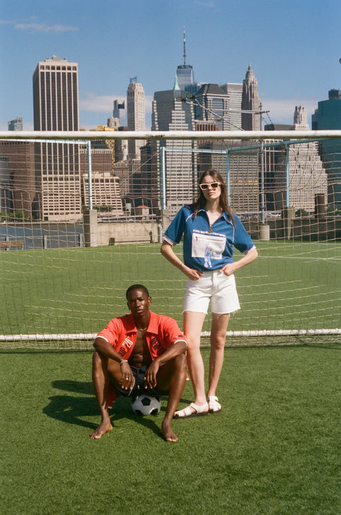 Two models wearing FIFA collection tops pose on a soccer field in front of a goal net with the New York City skyline behind them.