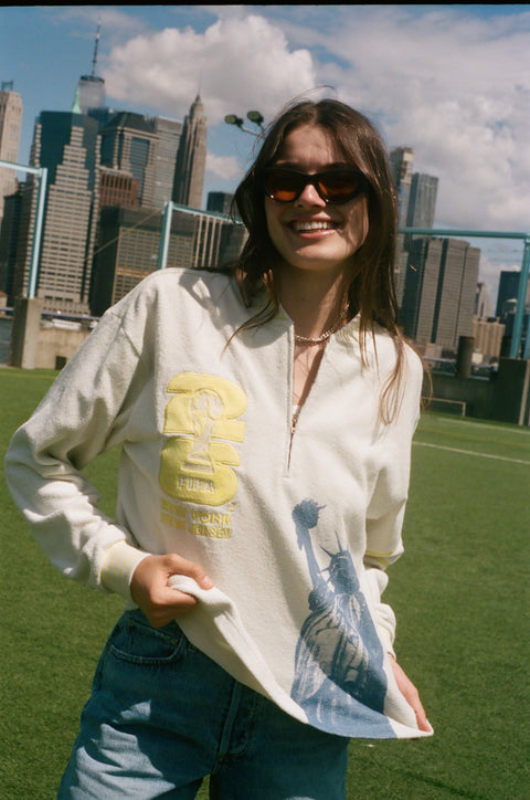 Model wearing a cream terry quarter-zip pullover with a Statue of Liberty graphic, standing outdoors with the New York City skyline behind her.