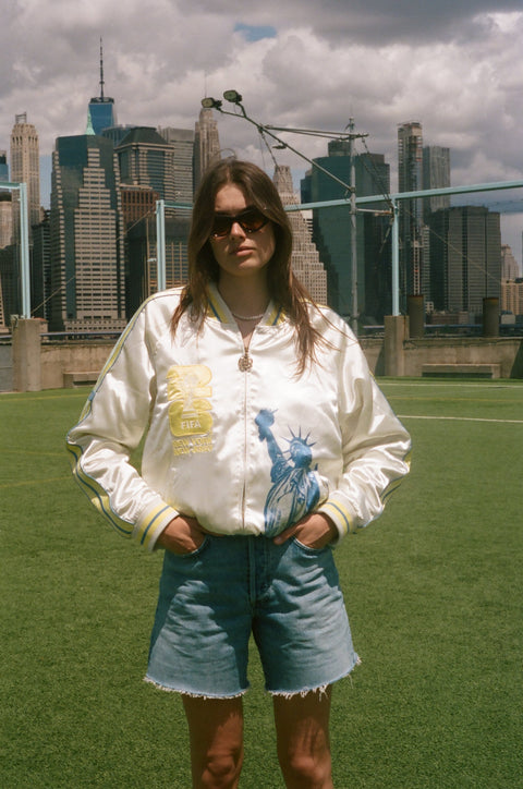Woman wearing the FIFA souvenir jacket with jean shorts, standing on a soccer field. 