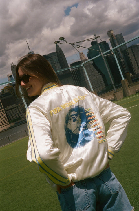 Person wearing a white jacket with graphics and text, standing outdoors with a cityscape in the background.