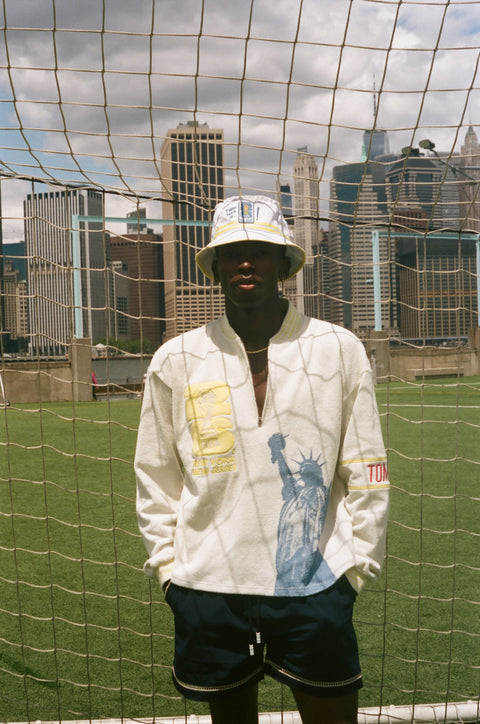 Model wearing a cream terry quarter-zip pullover with Statue of Liberty embroidery, standing in front of a soccer goal net.
