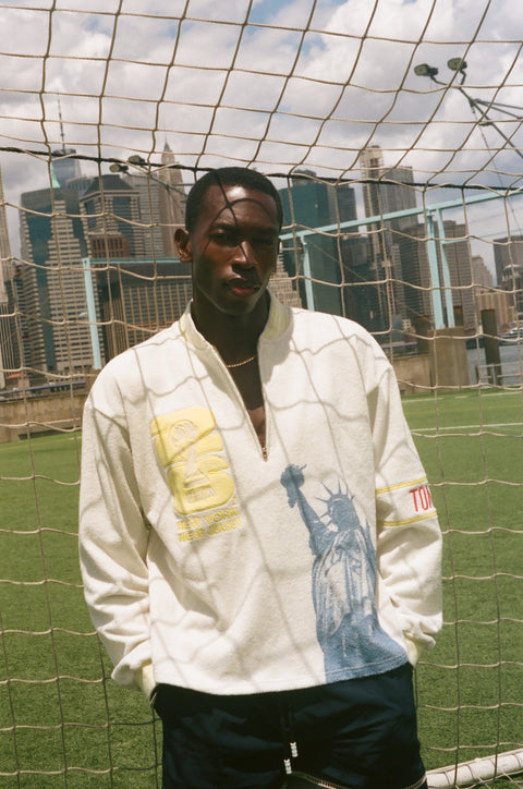 Model standing in front of a soccer goal on a waterfront field, wearing a cream terry quarter-zip pullover with pale yellow striped rib-knit trim, a yellow FIFA World Cup 26 New York/New Jersey crest on the chest, a blue Statue of Liberty graphic, and red Tombolo embroidery on the sleeve. Manhattan skyline visible in the background.