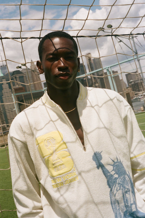 Close-up of a cream terry quarter-zip pullover with yellow FIFA embroidery and a blue Statue of Liberty graphic.