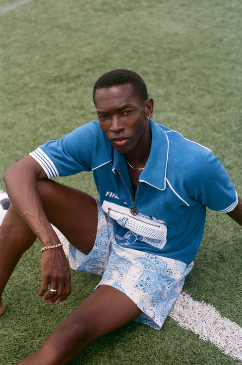 Model seated on a soccer field wearing a blue terry quarter-zip polo with FIFA World Cup embroidery and a mesh Statue of Liberty pocket.