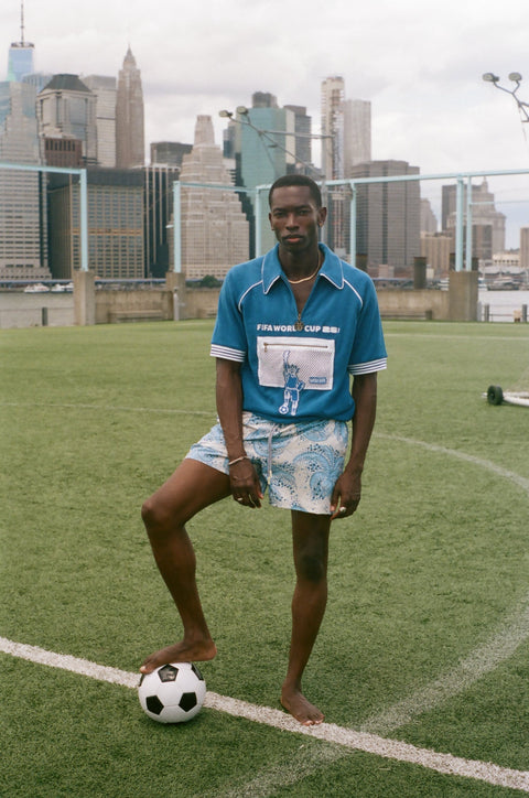 Model standing on a soccer field with a soccer ball, wearing a blue terry quarter-zip polo with FIFA World Cup embroidery and a mesh Statue of Liberty pocket.