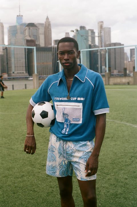Model wearing a blue terry quarter-zip polo with FIFA World Cup embroidery and a mesh Statue of Liberty pocket, holding a soccer ball on a grassy field.