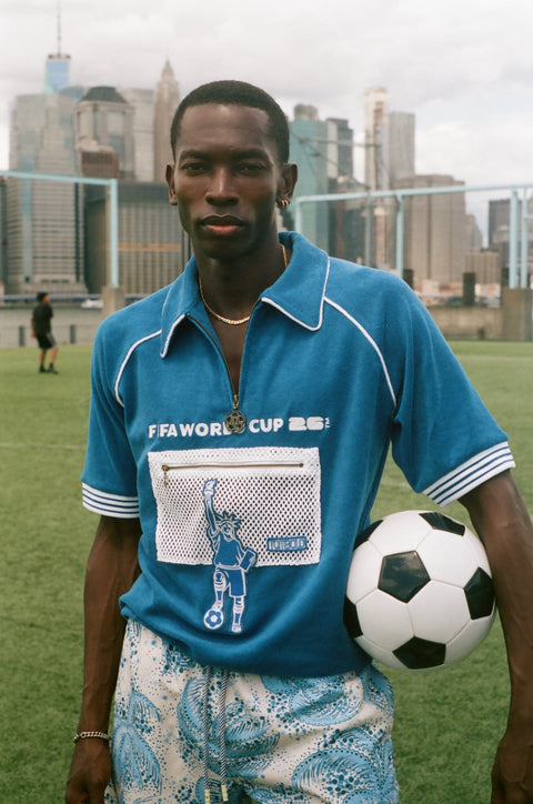 Model wearing a blue terry quarter-zip polo with FIFA World Cup embroidery and a mesh Statue of Liberty pocket, holding a soccer ball on a grassy field.