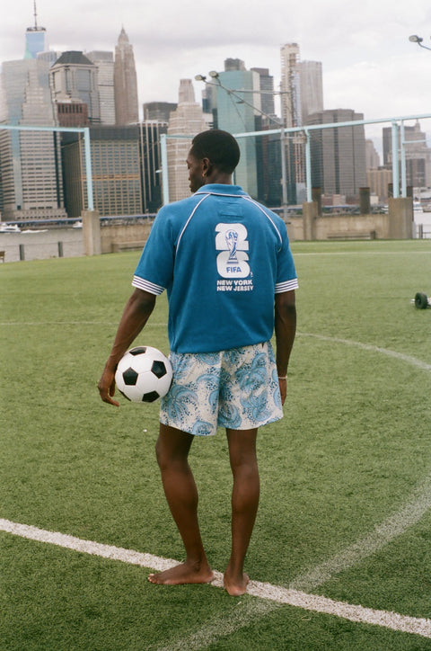 Back view of a blue terry quarter-zip polo with a large FIFA New York New Jersey graphic, standing on a soccer field with a soccer ball.