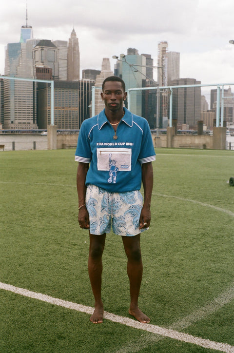 Full-length view of a model wearing a blue terry quarter-zip polo with FIFA World Cup embroidery and a mesh Statue of Liberty pocket, standing on a soccer field.