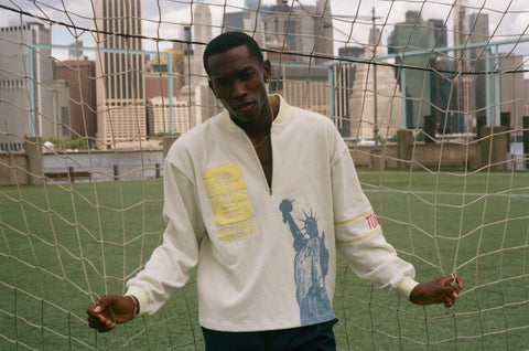 Model wearing a cream terry quarter-zip pullover with FIFA and Statue of Liberty graphics, standing in front of a soccer goal net.