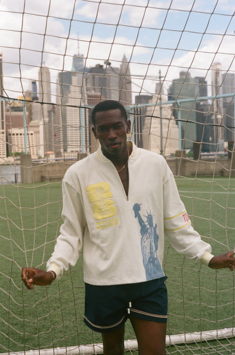 Model wearing a cream terry quarter-zip pullover with FIFA and Statue of Liberty graphics, standing in front of a soccer goal net.