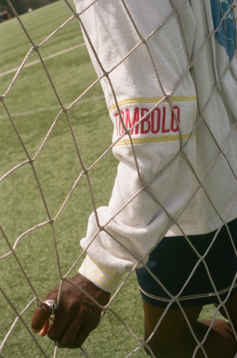 Close-up of a cream terry quarter-zip pullover sleeve with a red Tombolo logo patch behind a soccer goal net.