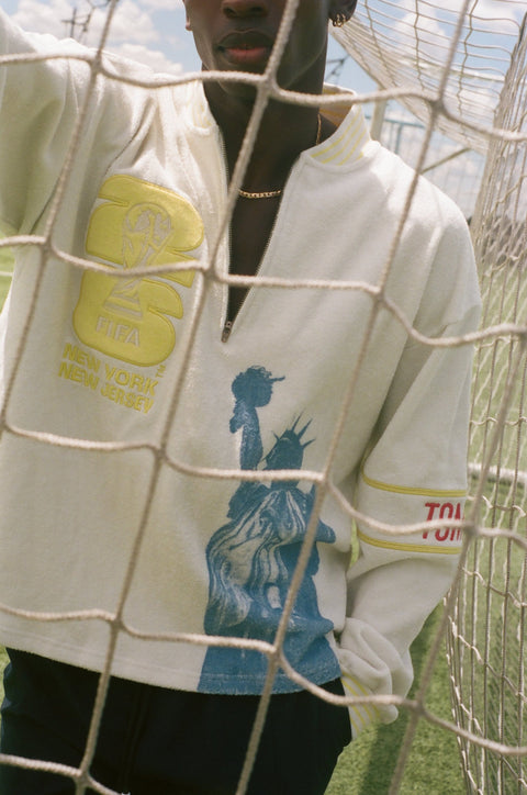 Close-up of a cream terry quarter-zip pullover with FIFA embroidery, a Statue of Liberty graphic, and a Tombolo logo sleeve patch behind a soccer goal net.