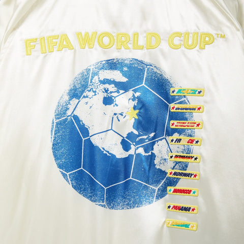 Close-up of the back artwork on a white satin bomber jacket, featuring yellow embroidered “FIFA WORLD CUP™” text, a blue globe graphic with a soccer ball pattern, a yellow star marking New York–New Jersey, and embroidered destination patches including Brazil, Ecuador, New York–New Jersey, France, Germany, Norway, Morocco, Panama, and Senegal.