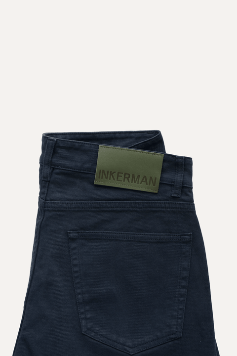 Close-up of the back waistband of navy jeans, showing a green rectangular brand patch with 'INKERMAN' printed on it - Inkerman NYC