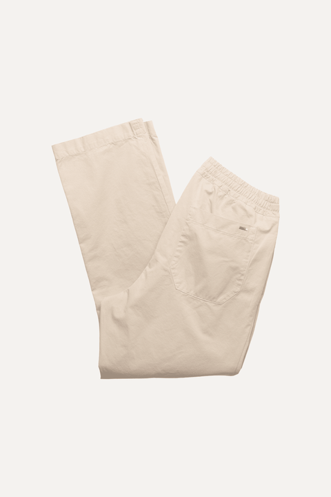 A pair of light beige trousers folded neatly on a solid off-white background, featuring an elastic waistband and a small embroidered logo on the side pocket - Inkerman NYC