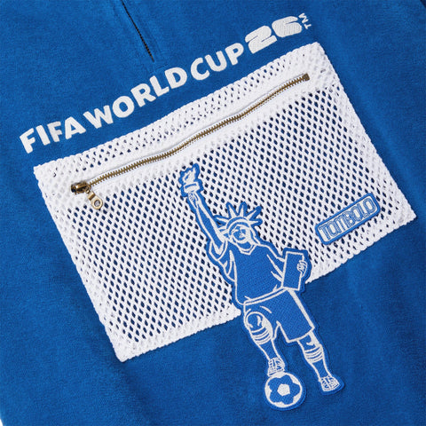 Close-up of a white mesh zip pocket with a Statue of Liberty soccer player embroidery and a Tombolo logo patch on a blue terry polo.