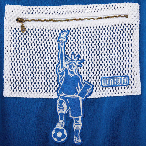 Close-up of a white mesh zip pocket with a Statue of Liberty soccer player embroidery and a Tombolo logo patch on a blue terry polo.